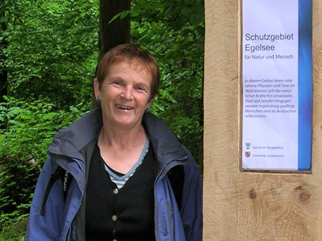 Das Bild zeigt ein Portrait von Susann Wehrli, welche sich im Wald an einen Markierungspfosten lehnt